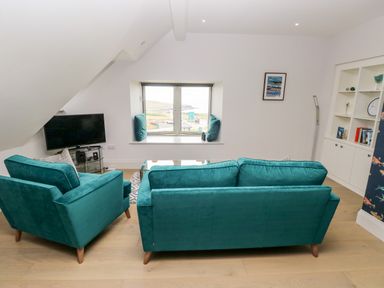A living room with teal sofa and armchair a glass table and TV near a window at 21 The Rest in Porthcawl