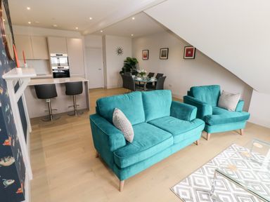 An open plan living room with teal sofas a glass coffee table a dining table with chairs and a kitchen area with stools at 21 The Rest in Porthcawl