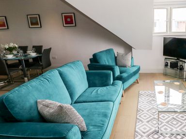 A living room with teal sofas, a glass coffee table on a patterned rug, a tv on a stand and a dining table with chairs and place settings at 21 The Rest in Porthcawl