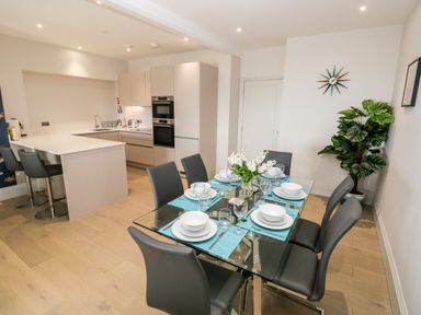 A dining area with a glass table set with plates and glasses next to a modern kitchen with bar stools and built-in appliances at 21 The Rest in Porthcawl