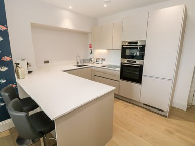 A modern kitchen with white countertops, beige cabinets, built-in ovens, a sink, and two black bar stools at 21 The Rest in Porthcawl