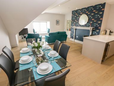 A dining area with a set table and teal placemats next to a living space with teal sofas and a window seat at 21 The Rest in Porthcawl