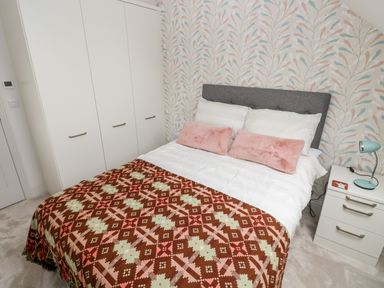 A bedroom with a double bed two pink cushions a patterned blanket a white wardrobe and a bedside table with a lamp at 21 The Rest in Porthcawl