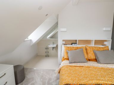 A bedroom with a bed with orange and gray pillows a small sink under a sloped ceiling and a gray ottoman at 21 The Rest in Porthcawl