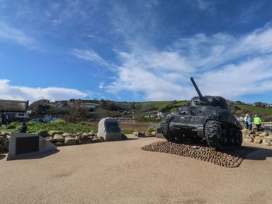 An outdoor memorial display featuring a tank and plaques with hills and houses in the background at Wrens Nest in Slapton