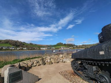An outdoor memorial with a tank and plaque next to a body of water and houses on distant hills at Wrens Nest in Slapton