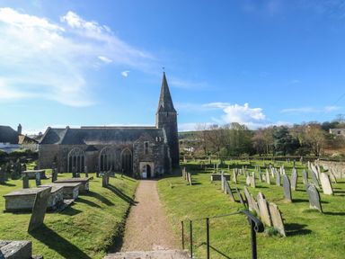 A church with a tall spire surrounded by a graveyard with numerous tombstones and a pathway at Wrens Nest in Slapton