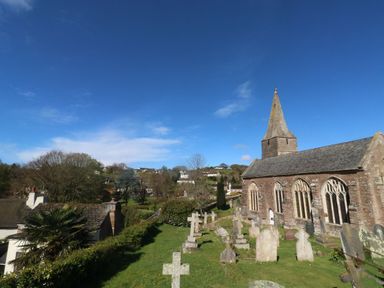 A church with a steeple and a graveyard with tombstones on a grassy area surrounded by trees and houses at Wrens Nest in Slapton