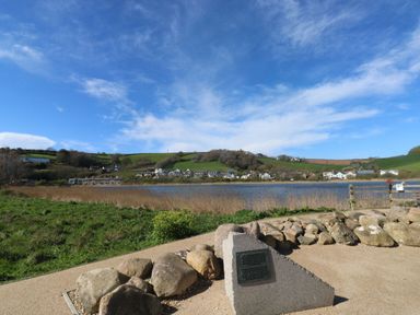 A lakeside view with a stone memorial plaque grassy bank and houses on distant hills at Wrens Nest in Slapton