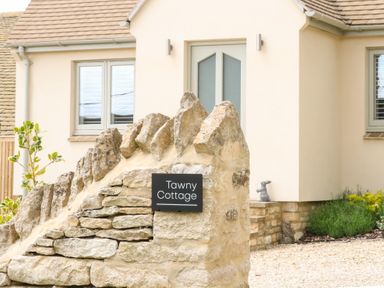 A stone wall with a sign reading Tawny Cottage outside a house with beige walls and grey framed windows at Tawny Cottage in Fulbrook near Burford
