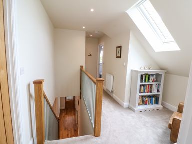 A hallway with carpeted floor and wooden staircase railing a white bookshelf under a skylight window and a framed picture on the wall at Tawny Cottage in Fulbrook near Burford