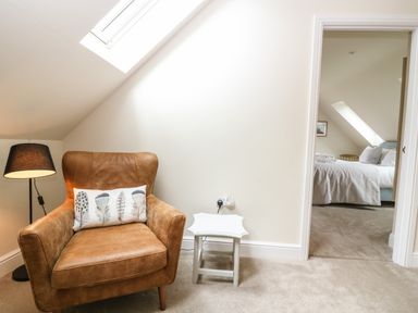 A brown leather armchair with a cushion next to a small white table and a floor lamp in a room with a carpet and a skylight at Tawny Cottage in Fulbrook near Burford
