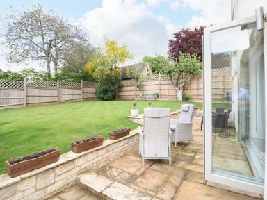 A backyard with a patio and white wicker chairs overlooking a lawn with trees and wooden fence at Tawny Cottage in Fulbrook near Burford