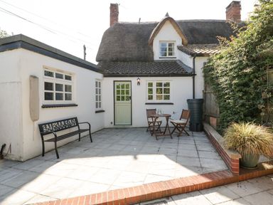 An outdoor patio area with a black bench two wooden chairs and a table beside a white building with a thatched roof at 1 Old Thatch in Kilve