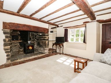 A living room with a stone fireplace wood-burning stove wooden beams on ceiling beige carpet and a window at 1 Old Thatch Kilve