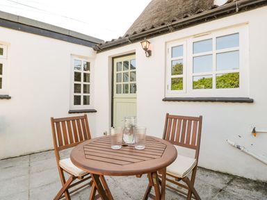 An outdoor patio with a round wooden table and two wooden chairs in front of a white building with windows and a door at 1 Old Thatch in Kilve
