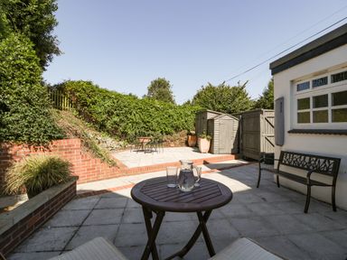 An outdoor patio area with a round wooden table and chairs a black metal bench wooden garden shed and green plants at 1 Old Thatch in Kilve