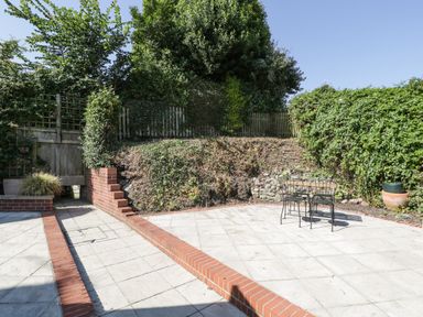 An outdoor patio area with a brick border table and chairs on a tiled floor surrounded by greenery and a small set of steps at 1 Old Thatch in Kilve