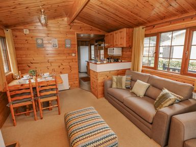 A wooden cabin interior with a dining table set for four a gray sofa with pillows a patterned ottoman and a kitchen area at Heritage Park Stepaside near Kilgetty