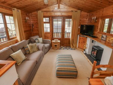 A living room with wooden walls and ceiling containing a sofa, armchair, patterned ottoman, television, and fireplace at Heritage Park Stepaside near Kilgetty