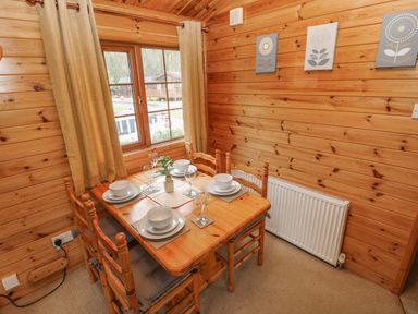 A dining area with a wooden table set for four near a window with beige curtains in a wooden cabin at Heritage Park in Stepaside near Kilgetty