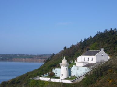 A white lighthouse and adjacent white house on a hillside overlooking a body of water at Apartment One in Duncannon County Wexford