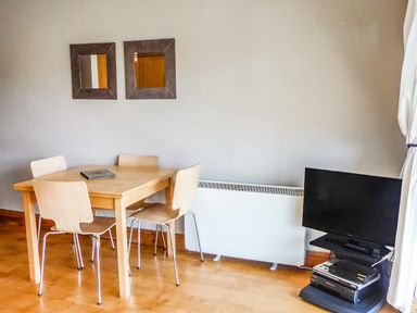 A dining area with a wooden table and four chairs next to a wall radiator and a television on a stand at Apartment One in Duncannon County Wexford
