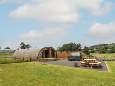 A curved wooden cabin with windows beside a hot tub and wooden picnic tables on a paved area in a grassy outdoor space at Bluebell in St Harmon near Rhayader