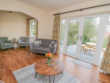 A living room with a round wooden coffee table with flowers on it and grey sofas near glass doors opening to a deck with a picnic table at Ger Yr Afon Cottage in St Harmon near Rhayader