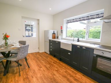 A kitchen with black cabinets a white countertop a white sink a dining table with grey chairs and a bouquet of flowers at Ger Yr Afon Cottage St Harmon near Rhayader