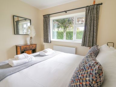 A bedroom with a bed, patterned cushions, folded towels on the bed, a wooden cabinet with a vase, a mirror on the wall, and a window with curtains at Ger Yr Afon Cottage in St Harmon near Rhayader