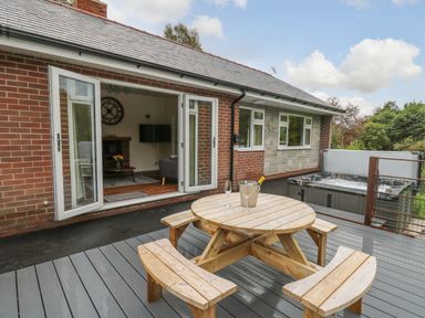 An outdoor patio with a wooden picnic table and benches and a hot tub next to a brick house with open doors at Ger Yr Afon Cottage in St Harmon near Rhayader