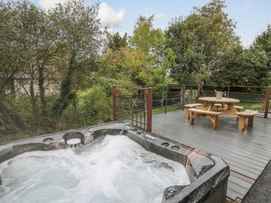An outdoor deck with a bubbling hot tub and a wooden picnic table with benches surrounded by trees at Ger Yr Afon Cottage St Harmon near Rhayader