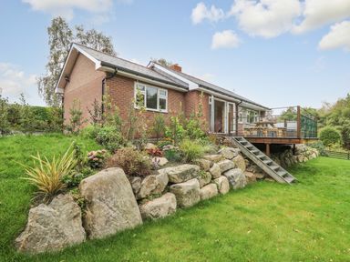 A brick house with a raised wooden deck stairs and landscaping with large rocks and plants at Ger Yr Afon Cottage in St Harmon near Rhayader