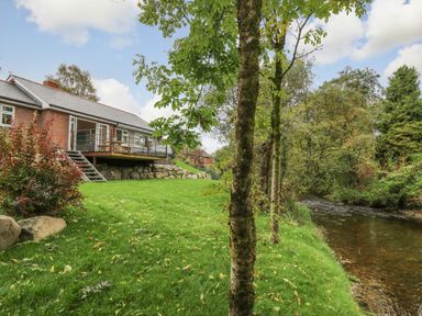 A house with a deck overlooking a river with trees and grass at Ger Yr Afon Cottage in St Harmon near Rhayader