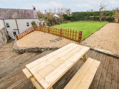 A wooden picnic table on a deck overlooking a garden with grass gravel and wooden fencing at The Old Bakery in Milton Tenby