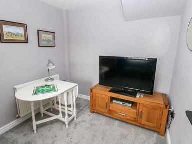 A small room with a television on a wooden stand and a white table with a lamp and a board game at The Old Bakery in Milton Tenby