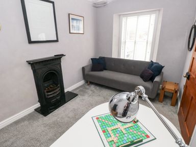 A room with a gray sofa and dark cushions next to a window with white blinds a black fireplace and a white table with a lamp and a scrabble board at The Old Bakery in Milton Tenby