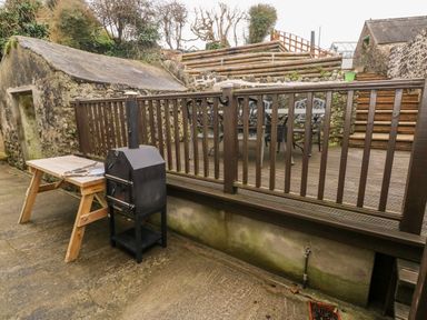 An outdoor patio area with a wooden bench table a black smoker grill and a raised wooden deck with a table and chairs at The Old Bakery in Milton Tenby