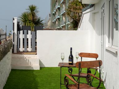 An outdoor patio with a wooden table and chairs a bottle and two glasses on the table and a white wall with a gate behind at Fistral Drift in Newquay