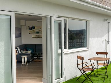 A small outdoor seating area with wooden table and chairs and an open door leading to a living room with a gray sectional sofa at Fistral Drift in Newquay