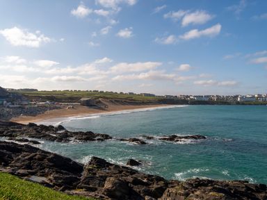 A coastline with rocky foreground a sandy beach green fields and buildings in the distance at Fistral Drift in Newquay
