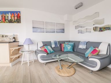 A living room with a curved sofa with colorful cushions a glass coffee table a side table with a lamp and wall art at Fistral Drift in Newquay