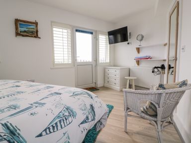A bedroom with a bed a white wicker chair a dresser a wall-mounted TV a door and windows with shutters at Fistral Drift in Newquay