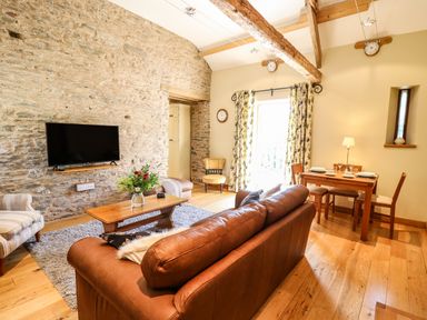 A living room with a brown leather sofa a wooden coffee table flowers on the table a wall mounted tv and a dining table set for four at Golwg Las in Pumsaint near Lampeter