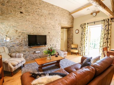 A living room with a stone wall TV a brown leather sofa a wooden coffee table and floral curtains at Golwg Las Pumsaint near Lampeter