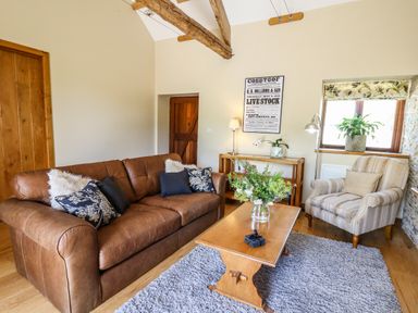 A living room with a brown leather sofa and striped armchair with cushions a wooden table with flowers and a wooden side table with a lamp and a framed poster on the wall at Golwg Las in Pumsaint near Lampeter