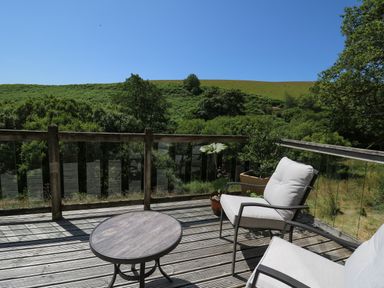 An outdoor wooden deck with two cushioned chairs a round table and potted plants overlooking green hills at Golwg Las in Pumsaint near Lampeter