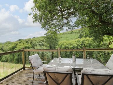 An outdoor patio with a glass table and four cushioned chairs overlooking green trees and hills at Nant Glas in Pumpsaint near Lampeter