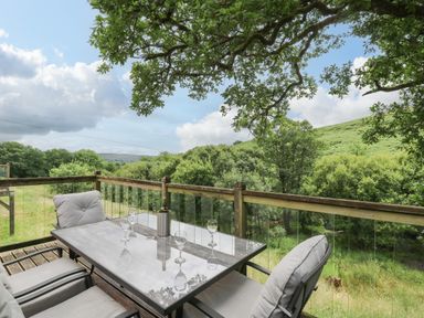 An outdoor patio with a glass table and cushioned chairs overlooking trees and hills at Nant Glas Pumpsaint near Lampeter